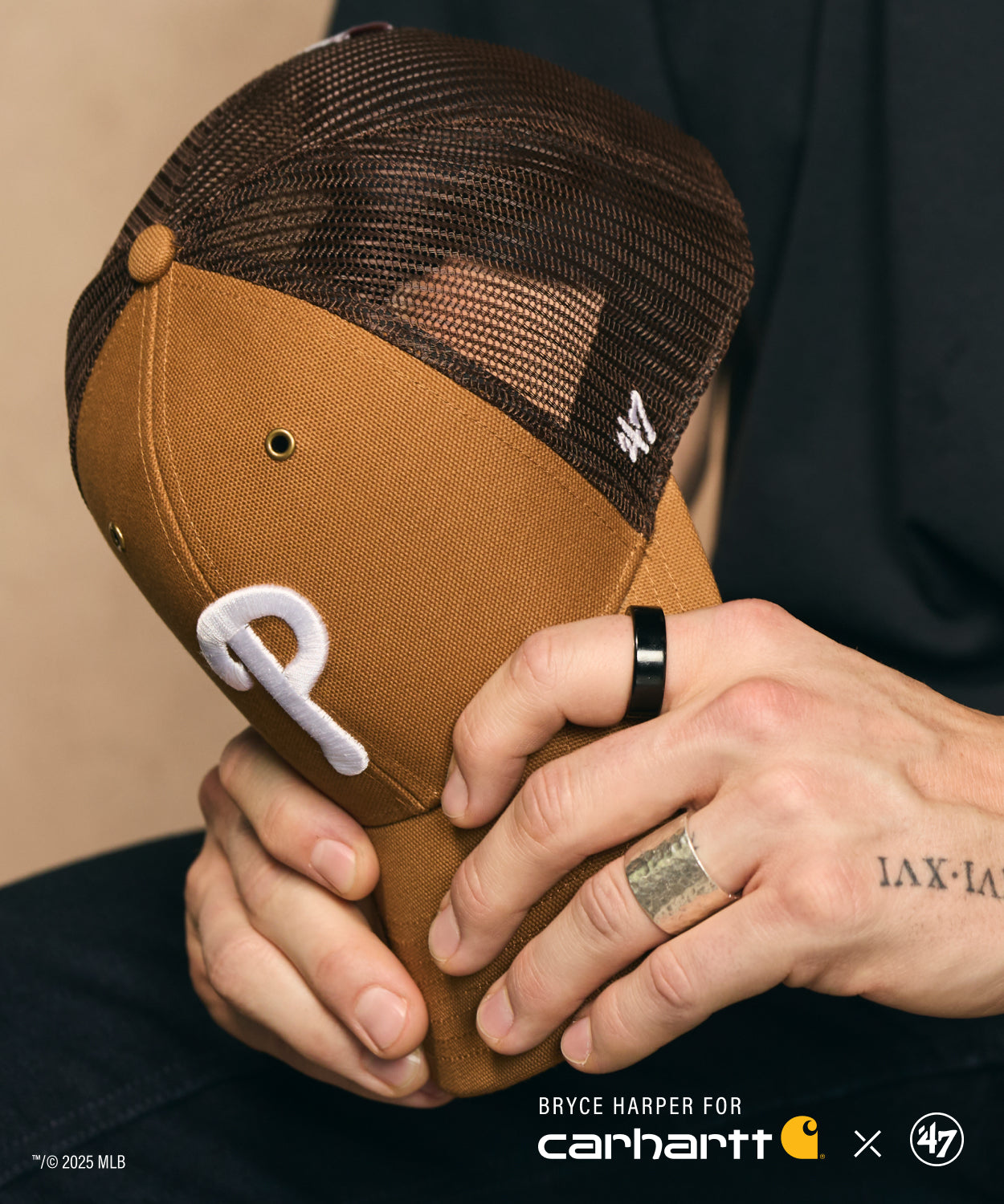 Person holding a brown '47 CARHARTT baseball cap with a logo, wearing a black shirt.